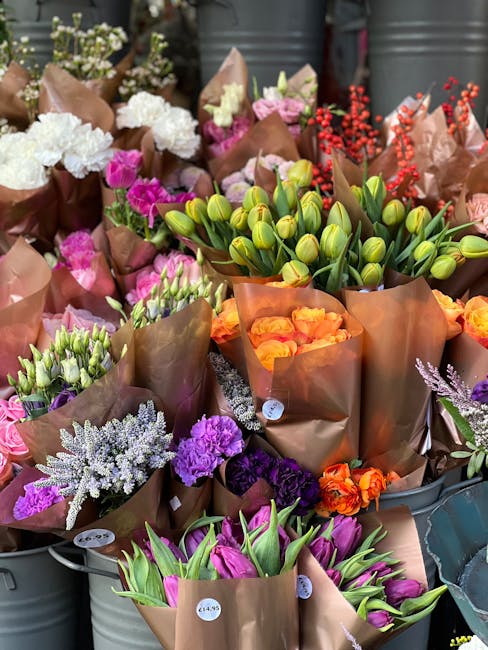 A vibrant display of fresh floral bouquets arranged in metal buckets and wrapped in brown paper, featuring a variety of flowers including white hydrangeas, pink and purple carnations, orange roses, pu