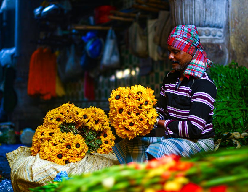 A male flower vendor sitting outdoors in a vibrant market setting, holding a bouquet of bright yellow sunflowers with dark centers. Several large sunflower bouquets are displayed on a woven cloth on t