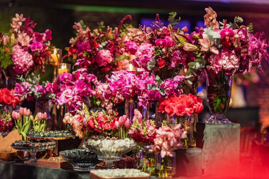 A vibrant arrangement of fresh pink and red flowers, including orchids, roses, tulips, and hydrangeas, displayed in glass vases and bowls on a dark table surface. The flowers are arranged in various h