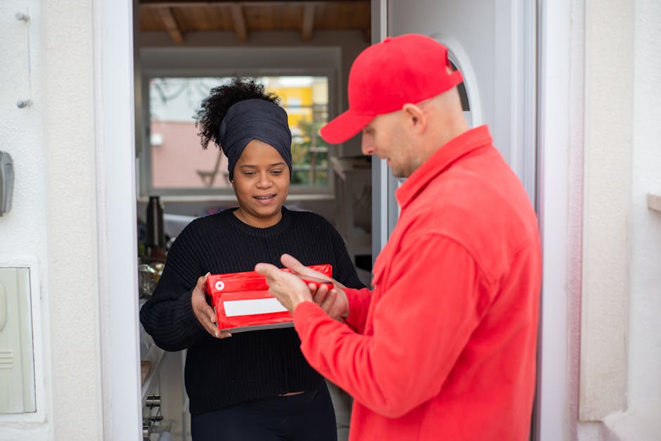 A delivery person dressed in a red cap and jacket is handing over a red parcel to a woman at her doorway. The woman, with dark hair styled in a headscarf, is holding a bouquet arrangement of fresh, vi