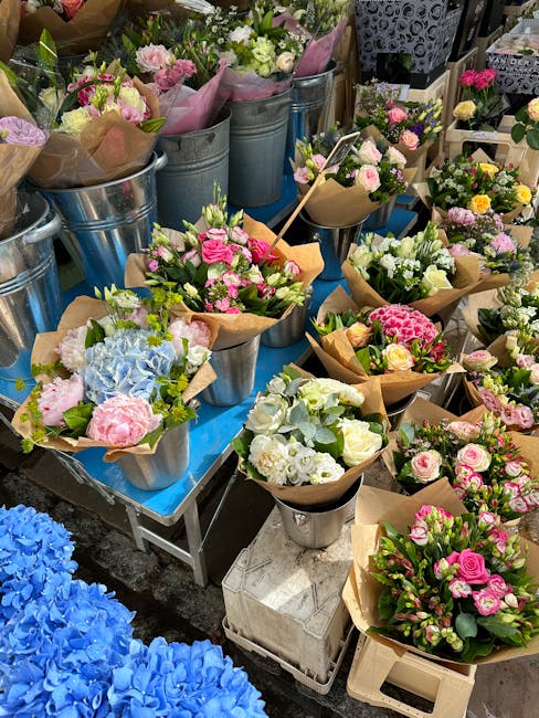 An outdoor display of fresh floral bouquets arranged in metal buckets and wrapped in brown paper, featuring pink, white, and pastel-colored roses, hydrangeas, and other blooms. The bouquets are placed