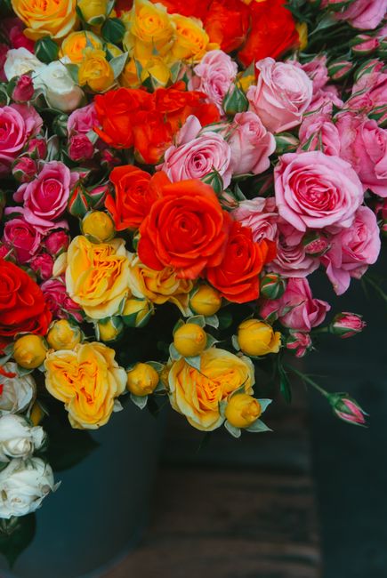 A vibrant floral arrangement featuring a mix of roses in various shades of pink, red, yellow, and orange. The roses are densely clustered, showcasing their layered petals with some still in bud form a